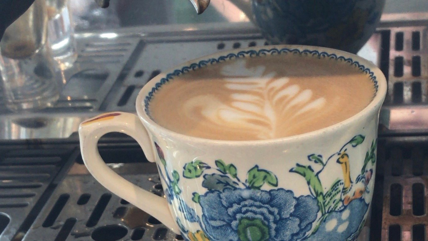 A flat white with leaf-pattern latte art in the café's vintage blue-and-white floral cup, on a La Marzocco espresso machine